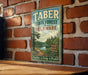 A vintage metal sign for the Taber State Forest in Delaware, featuring a scenic image of a forest and a lake, mounted on a brick wall.