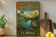 A vintage-style metal sign for Tate's Hell State Forest in Florida, featuring a tropical landscape with palm trees, a lake, and an alligator.