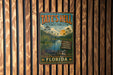 A vintage metal sign for Tate's Hell State Forest in Florida, featuring a tropical landscape with palm trees, a crocodile, and a sunset.