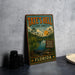 A vintage-style metal sign for Tate's Hell State Forest in Florida, featuring a tropical landscape with palm trees, a body of water, and an alligator.