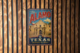 A vintage-style metal sign with the text "The Alamo" and "Texas Remember the Alamo!" displayed on a wooden wall.