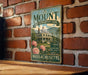 A vintage-style metal sign for "The Mount Edith Wharton's Home" in Massachusetts, featuring a large illustration of a house surrounded by flowers and a mountainous landscape.