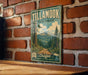 A vintage-style metal sign for Tillamook State Forest, featuring a scenic mountain landscape and trees, mounted on a brick wall.