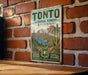 A vintage-style metal sign for Tonto National Forest in Arizona, featuring a desert landscape with mountains, cacti, and a body of water.