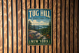 A vintage metal sign for Tug Hill State Forest in New York, featuring a scenic landscape with pine trees, a lake, and mountains.