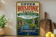A vintage-style sign for the Upper Housatonic Valley National Heritage Area in Connecticut, featuring a scenic illustration of a river, buildings, and a person standing on a cliff overlooking the water.