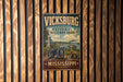 A vintage metal sign for Vickburg National Military Park in Mississippi, featuring a cannon and a river, mounted on a wooden wall.
