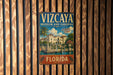 A vintage metal sign for the Vizzaya Museum and Gardens in Florida, featuring a tropical scene with palm trees and a building.
