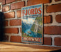 A vintage travel poster advertising West Norwegian Fjords, featuring a scenic image of a waterfall and mountains, is displayed on a brick wall.