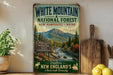A vintage-style sign for White Mountain National Forest in New Hampshire, Maine, featuring a scenic mountain landscape with a river and pine trees.