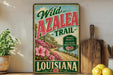 A vintage-style metal sign advertising the Wild Azalea Trail in Louisiana, featuring a scenic landscape with a road, flowers, and a river.