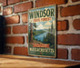 A vintage metal sign for the Windsor State Forest in Massachusetts, featuring a scenic landscape with pine trees and a lake.