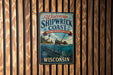 A vintage-style metal sign for the Wisconsin Shipwreck Coast, featuring an illustration of a shipwreck and a lighthouse, mounted on a wooden wall.