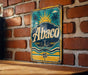 A vintage Abaco sign is displayed on a brick wall, featuring a sun and palm tree design.