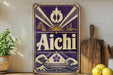 a vintage-style sign with the word "Aichi" prominently displayed, surrounded by various decorative elements such as a sunburst design, waves, and a temple. The sign is placed on a wooden surface, with a potted plant and a wooden cutting board visible in the background.