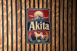 A vintage-style metal sign with the word "Akita" prominently displayed, featuring a dog and a mountain landscape.