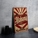 A vintage-style metal sign with the word "Alaska" prominently displayed, featuring a sunburst design and a small building. The sign is placed on a gray surface, with a stack of black camera lenses and a white dish visible in the background.