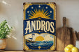 A vintage-style metal sign with the word "ANDROS" prominently displayed, featuring a lighthouse and a sun-like design. The sign is placed on a wooden surface, with a potted plant, a wooden cutting board, and some lemons nearby.