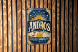 A vintage-style sign with the word "ANDROS" prominently displayed, featuring a lighthouse and a sun-like symbol.