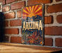 A vintage-style metal sign with the word "ARIZONA" prominently displayed, featuring a desert landscape with mountains and a sunburst design.