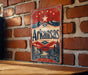 A vintage-style sign for Arkansas, featuring a red, white, and blue design with stars and mountains, mounted on a brick wall.