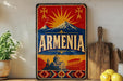 a vintage-style sign with the word "ARMENIA" prominently displayed, along with a mountain and sun motif. The sign is placed on a shelf next to a wooden cutting board and a potted plant.