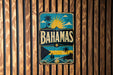 A vintage-style metal sign with the word "BAHAMAS" prominently displayed, featuring a tropical beach scene with palm trees, a sun, and a boat.
