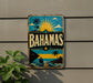 A vintage-style sign with the word "BAHAMAS" prominently displayed, featuring a tropical beach scene with palm trees, a sun, and a sailboat.