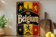 A vintage-style metal sign with the word "Belgium" prominently displayed, featuring a coat of arms and a castle-like structure. The sign is placed on a wooden surface, with a potted plant, a wooden cutting board, and some lemons nearby.