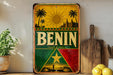 a vintage-style sign with the word "BENIN" prominently displayed, surrounded by a tropical landscape with palm trees and a sun. The sign is placed on a wooden surface, with a potted plant and a wooden cutting board nearby.