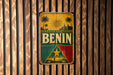 a vintage-style metal sign with the word "BENIN" prominently displayed, surrounded by a tropical landscape with palm trees and a sun.