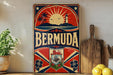 A vintage-style metal sign with the word "BERMUDA" prominently displayed, featuring a sun, lighthouse, palm trees, and other tropical elements. The sign is placed on a wooden surface, with a potted plant, a wooden cutting board, and some lemons nearby.