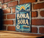 A vintage-style metal sign with the word "BORA" prominently displayed, featuring a tropical landscape with palm trees and a mountain in the background.