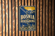 A vintage-style poster or sign with the text "Bosnia and Herzegovina" displayed prominently in the center, surrounded by a decorative border.