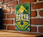 A vintage metal sign with the word "BRAZIL" prominently displayed, featuring a tropical landscape with palm trees and a sun.