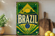A vintage-style sign with the word "BRAZIL" prominently displayed, featuring a tropical landscape with palm trees and a sun. The sign is placed on a shelf next to a wooden cutting board and a potted plant.