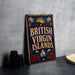 A vintage-style sign for the British Virgin Islands, featuring a sun and palm tree design, is displayed on a gray countertop next to a stack of black camera lenses.
