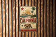 A vintage-style metal sign with the word "CALIFORNIA" prominently displayed, featuring a sun, palm trees, and a bear.