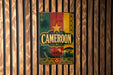 A vintage metal sign with the word "Cameroon" prominently displayed, featuring a colorful design with a palm tree, sun, and mountains.