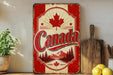 A vintage-style metal sign with the word "Canada" prominently displayed, featuring a maple leaf and mountain landscape. The sign is placed on a shelf next to a wooden cutting board and a potted plant.