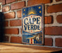 A vintage metal sign with the word "CAPE VERDE" displayed prominently, featuring a sun and stars design, is mounted on a brick wall.