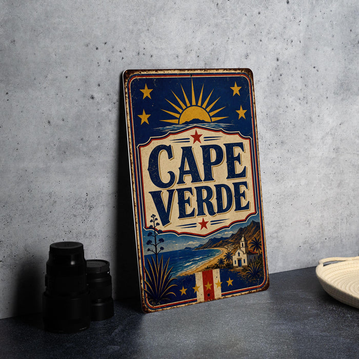 A vintage-style metal sign with the text "CAPE VERDE" displayed prominently, featuring a sun and stars design. The sign is placed on a gray surface, with a stack of black camera lenses and a white dish visible in the background.
