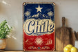 A vintage-style metal sign with the word "Chile" prominently displayed, featuring a star, palm trees, mountains, and a sun. The sign is placed on a shelf next to a wooden cutting board and some potted plants.