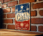 A vintage metal sign with the word "Chile" prominently displayed, featuring a sun and palm trees, against a brick wall background.