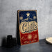 A vintage-style metal sign with the word "Chile" prominently displayed, featuring a red, white, and blue color scheme with a star and palm trees. The sign is placed on a gray surface, with a stack of black camera lenses and a white dish visible in the background.