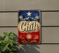 A vintage-style metal sign with the word "Chile" prominently displayed, featuring a star, palm trees, and mountains in the background.