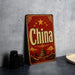 A vintage-style sign with the word "China" prominently displayed, featuring an illustration of a Chinese temple and mountains. The sign is placed on a gray surface, with some black camera lenses visible in the background.