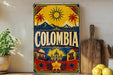 a vintage-style sign with the word "COLOMBIA" prominently displayed, surrounded by various decorative elements such as a sun, palm trees, mountains, and a building. The sign is placed on a shelf next to a wooden cutting board and a potted plant, creating a visually appealing and colorful display.