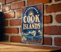 A vintage-style sign for Cook Islands, featuring a sailboat and tropical landscape, is displayed on a brick wall.