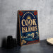 A vintage-style sign for Cook Islands, featuring a tropical island landscape with a sailboat, palm trees, and a sun.
